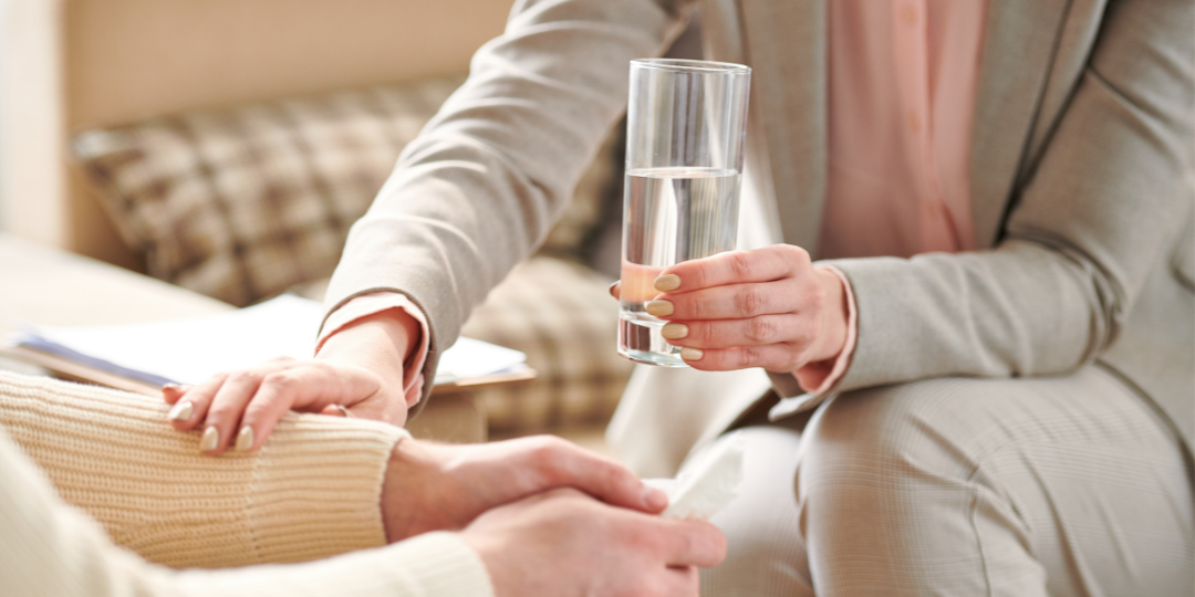 Close up, one woman has hand on another's arm and offers a glass of water