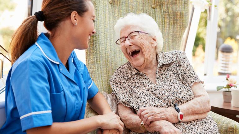 Seated elderly lady wearing Telecare wristband, laughing with female carer 